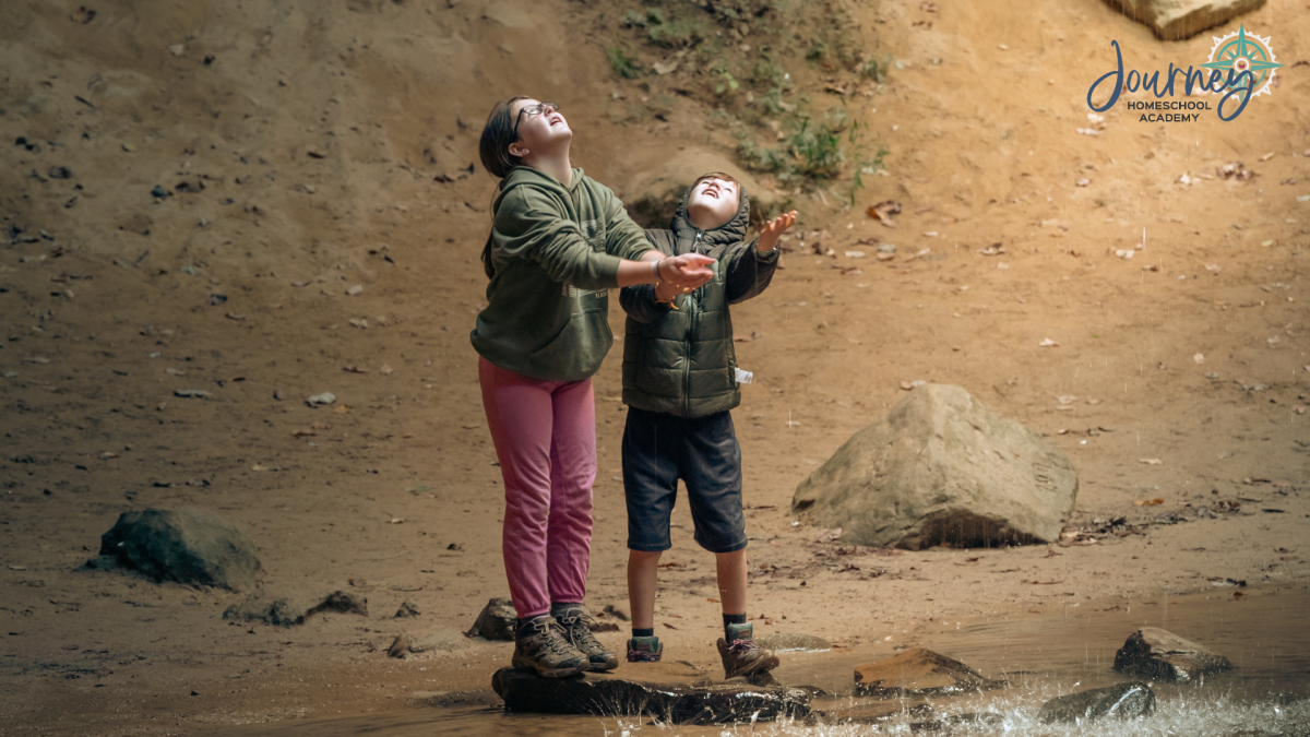 Two children in outdoor clothes reach up toward the rain while exploring nature during an earth science homeschool activity.