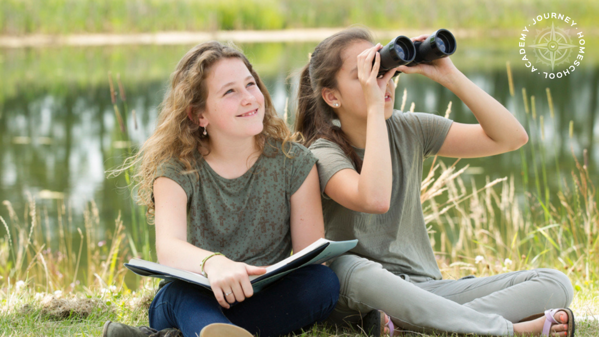Two girls using binoculars and nature journals by a pond during a bird nesting homeschool activity with Journey Homeschool Academy.